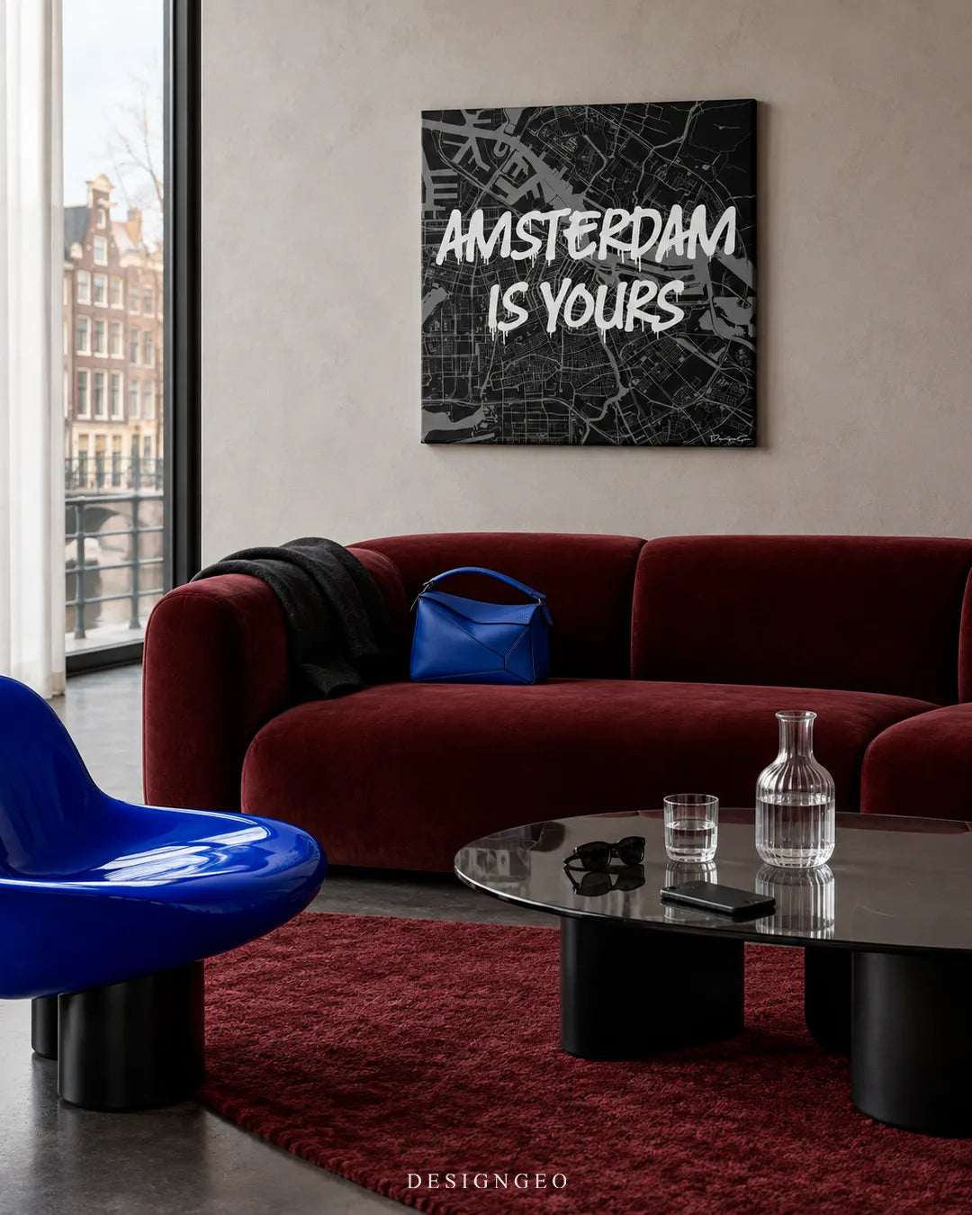 Modern luxury living room decor featuring a square canvas print of a black and white Amsterdam city map overlaid with white graffiti text AMSTERDAM IS YOURS above a deep red velvet sofa