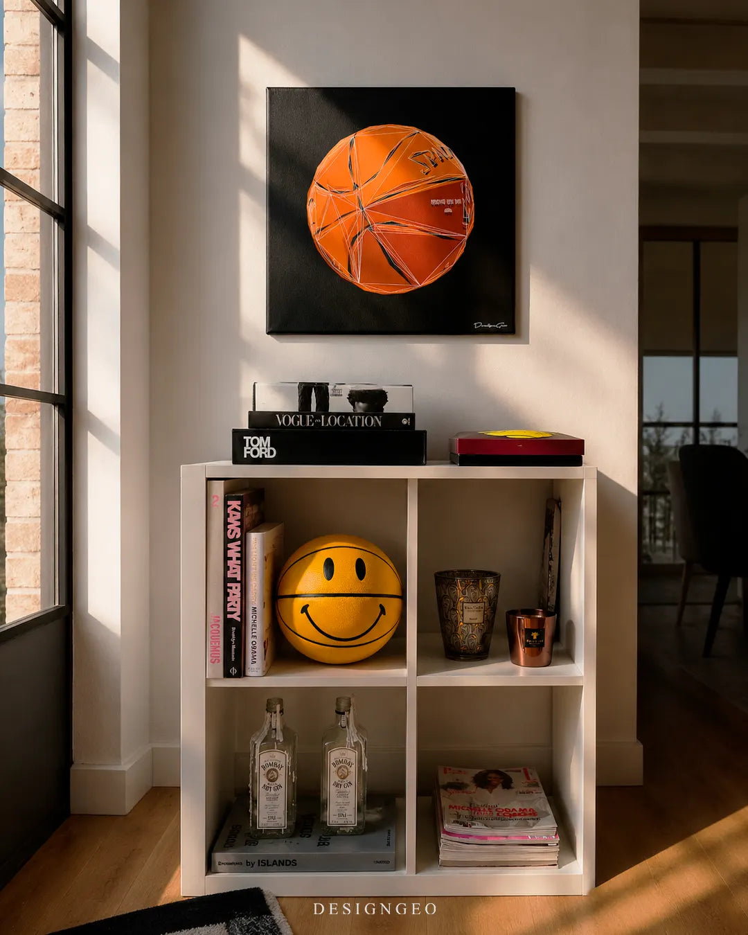 Square geometric basketball canvas print mounted on a white wall above a white cube shelving unit styled with a yellow smiley basketball, Tom Ford books, and Bombay Sapphire gin bottles
