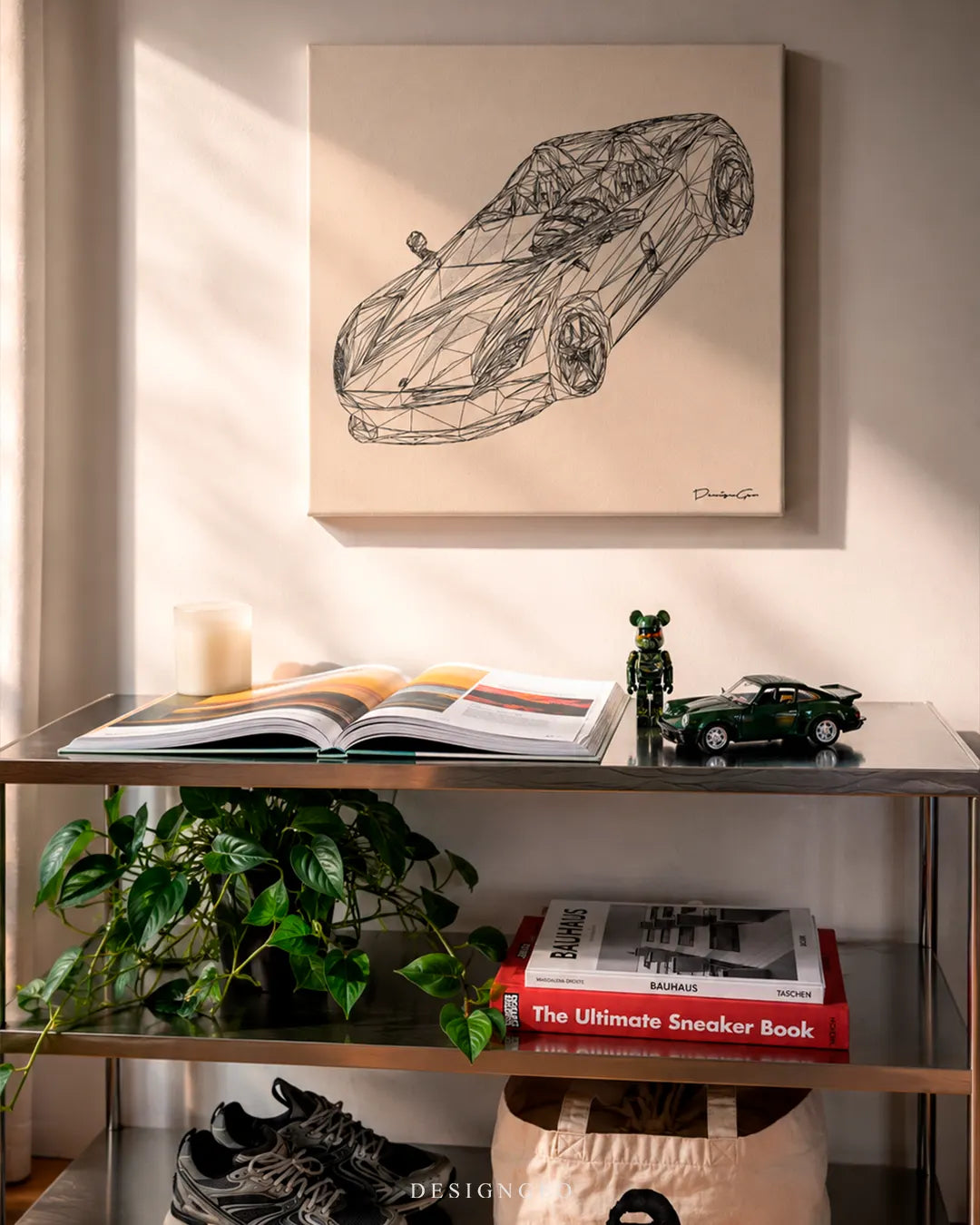 Square beige exotic supercar wireframe canvas print mounted on a wall above a stainless steel shelf unit with an open design book, green Porsche scale model car, Bearbrick figure, potted plant, and design books