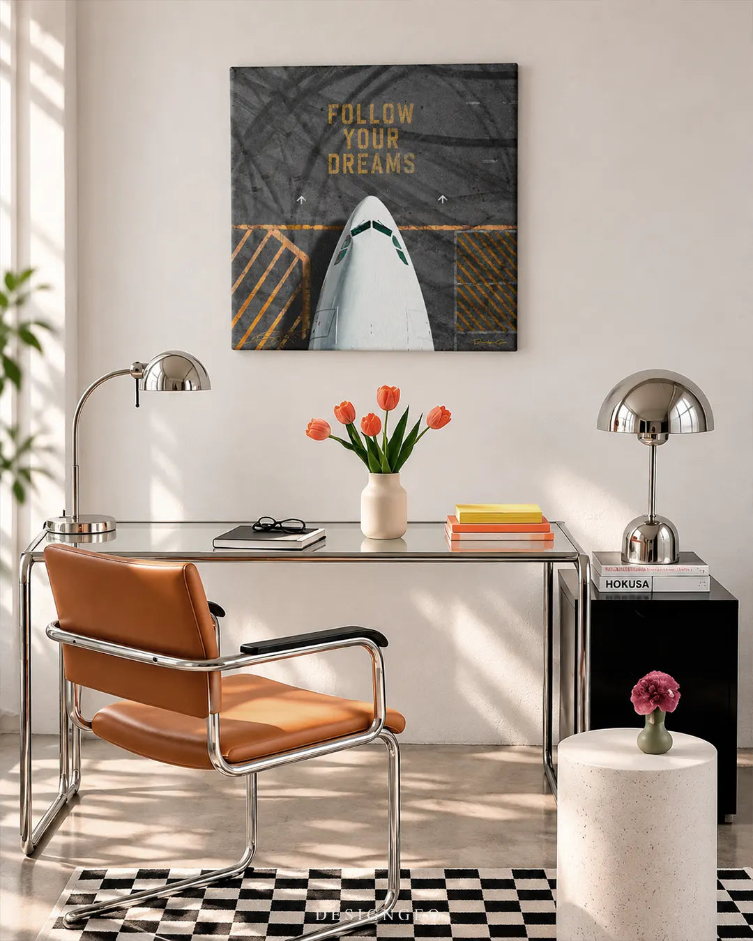 Square gray canvas print featuring the nose of a commercial airplane with gold typography spelling FOLLOW YOUR DREAMS mounted on a white wall above a modern glass desk with a brown leather chair