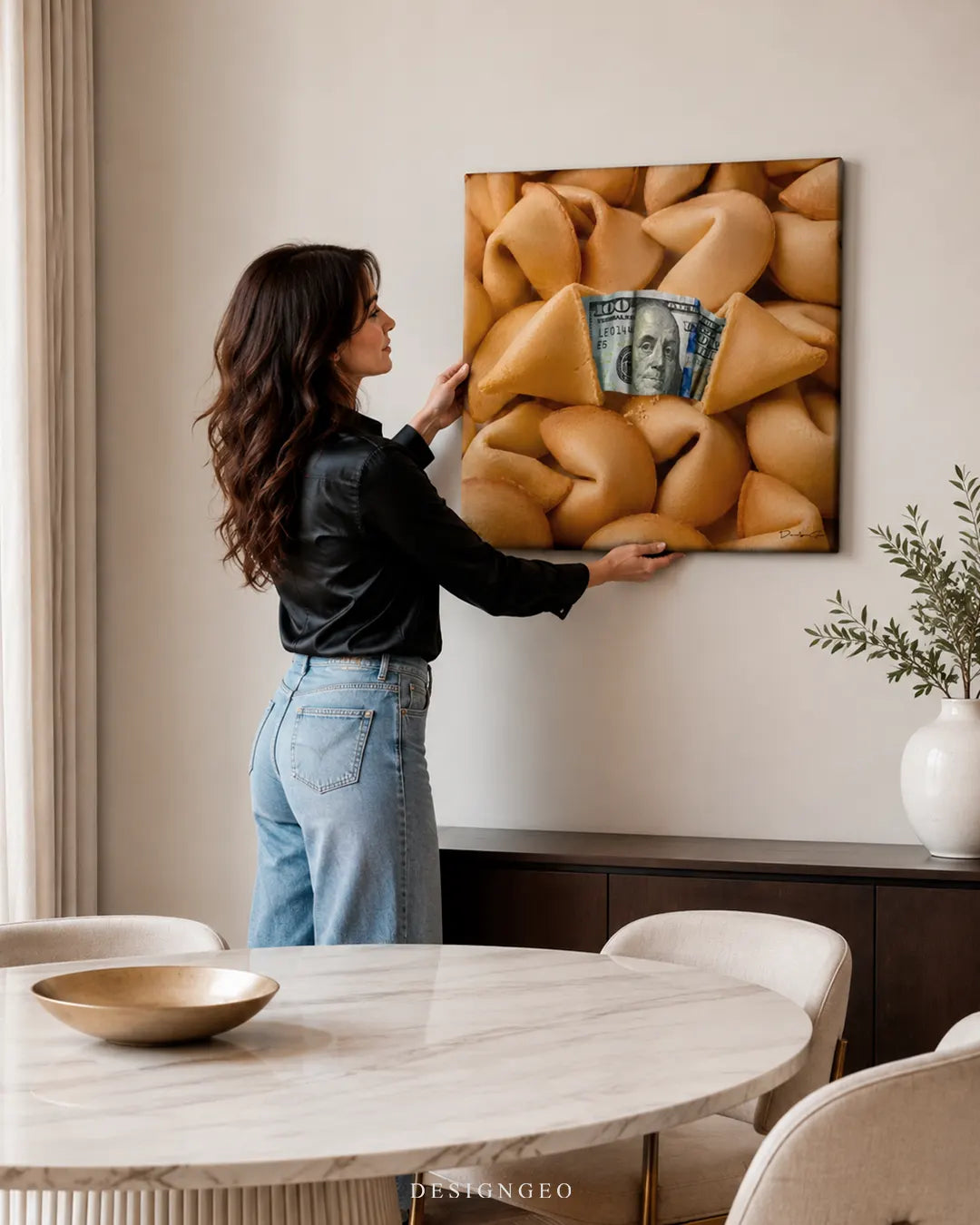 Woman hanging a square fortune cookie canvas art with a 100 dollar bill reveal on a modern dining room wall above a sideboard