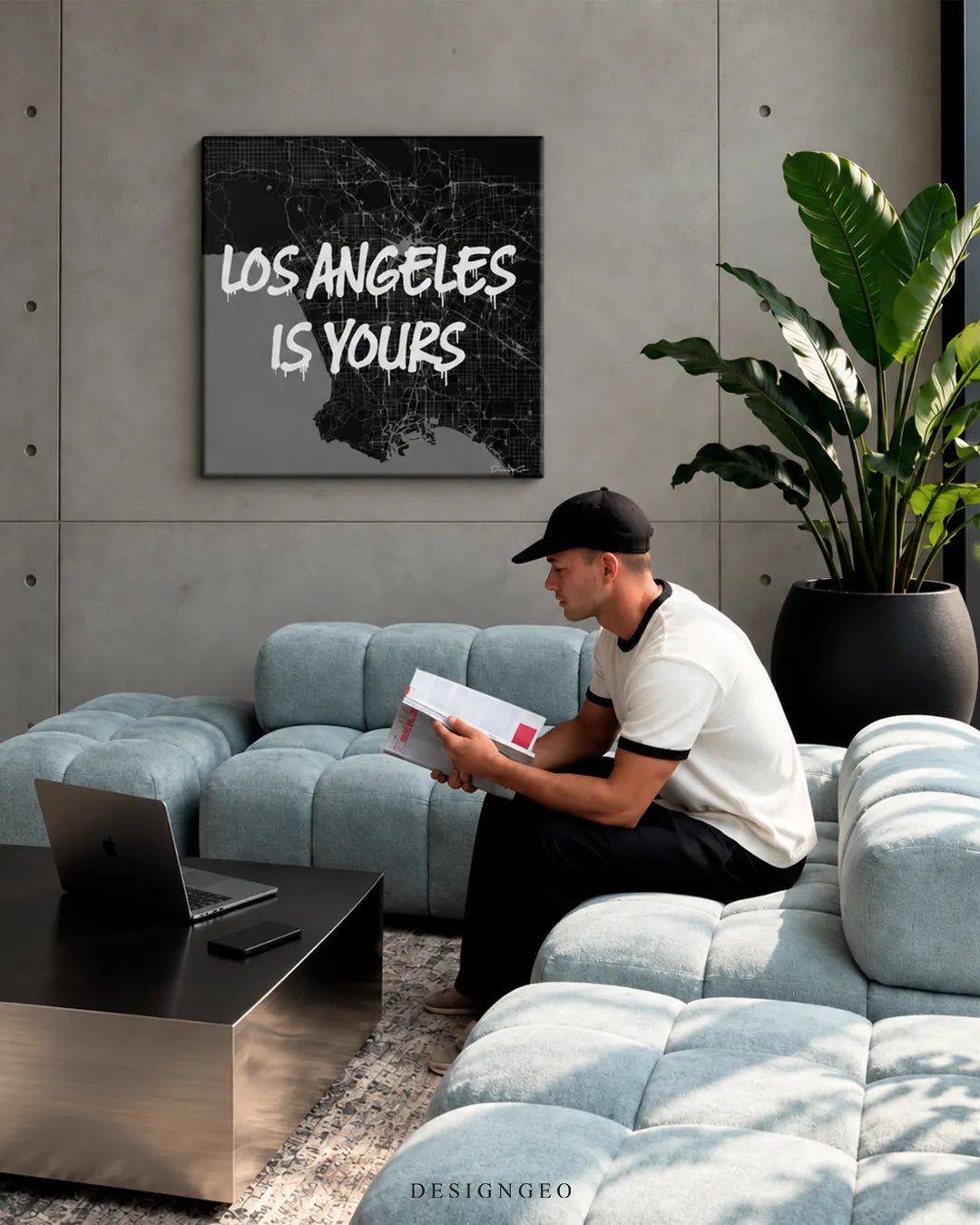 Los Angeles is Yours white spray paint text on black city map square canvas wall art displayed on a concrete wall in a modern living room above a light blue modular sofa with a person reading