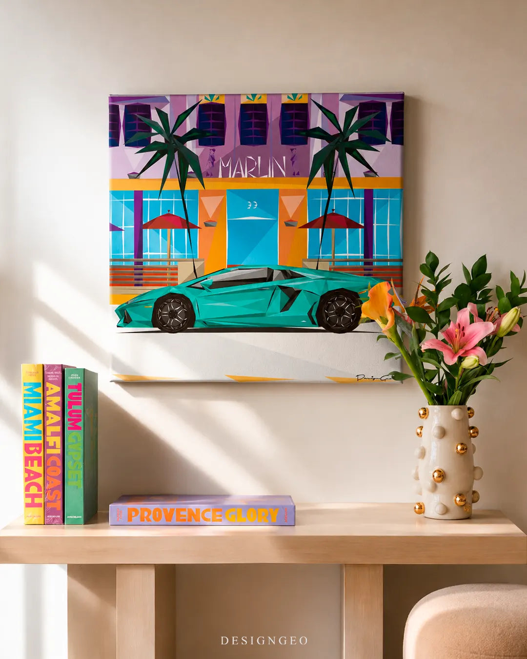 Square South Beach sports car canvas print mounted on a beige wall above a light wood console table with Assouline travel books and pink lilies in a studded vase