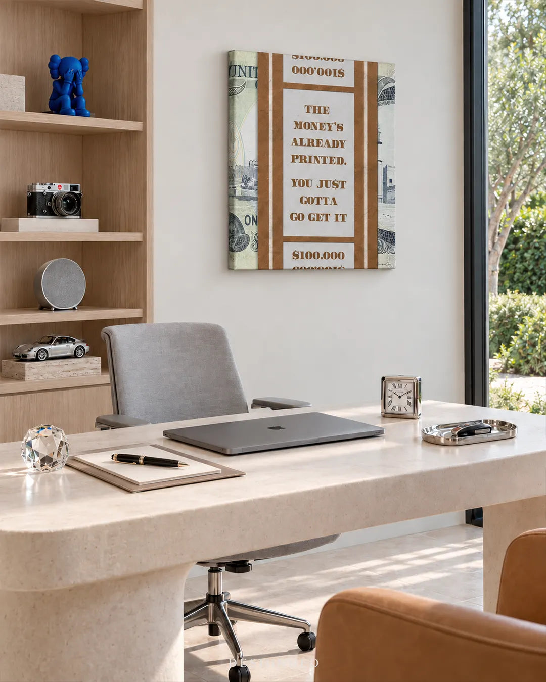 Square canvas print showing a brown AED banknote strap with motivational quote THE MONEY'S ALREADY PRINTED hanging on a white wall in an executive office above a beige desk