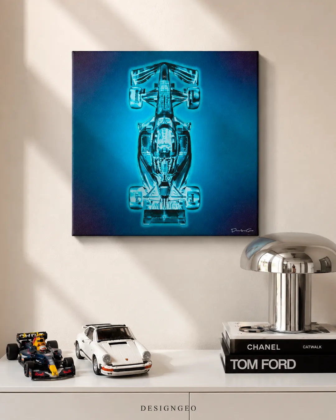 Square formula race car x-ray canvas print mounted on a beige wall above a white sideboard styled with a Red Bull F1 model car, classic white Porsche 911 scale model, Chanel book, Tom Ford book, and silver dome table lamp