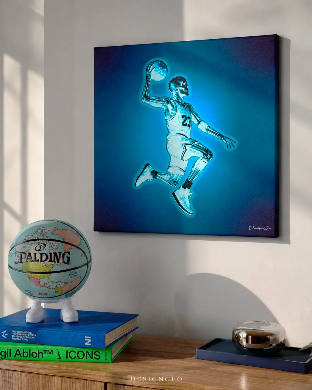 Square basketball anatomy x-ray canvas print mounted on a white wall above a wooden credenza with a globe Spalding basketball, Virgil Abloh ICONS book, and navy tray