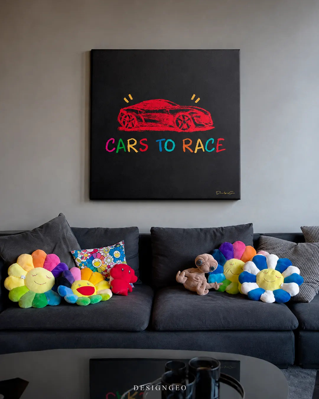 Square black Cars to Race canvas print mounted on a modern grey wall above a charcoal sofa decorated with colorful Takashi Murakami flower plush pillows