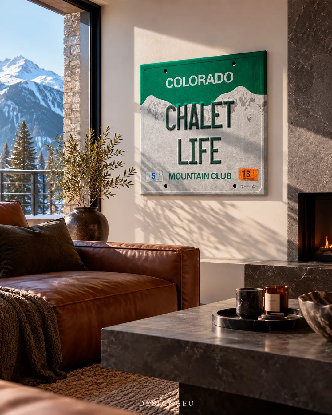Rustic modern ski lodge living room with snow-capped mountain views featuring a square Colorado Chalet Life license plate canvas print mounted above a brown leather sofa near a stone fireplace