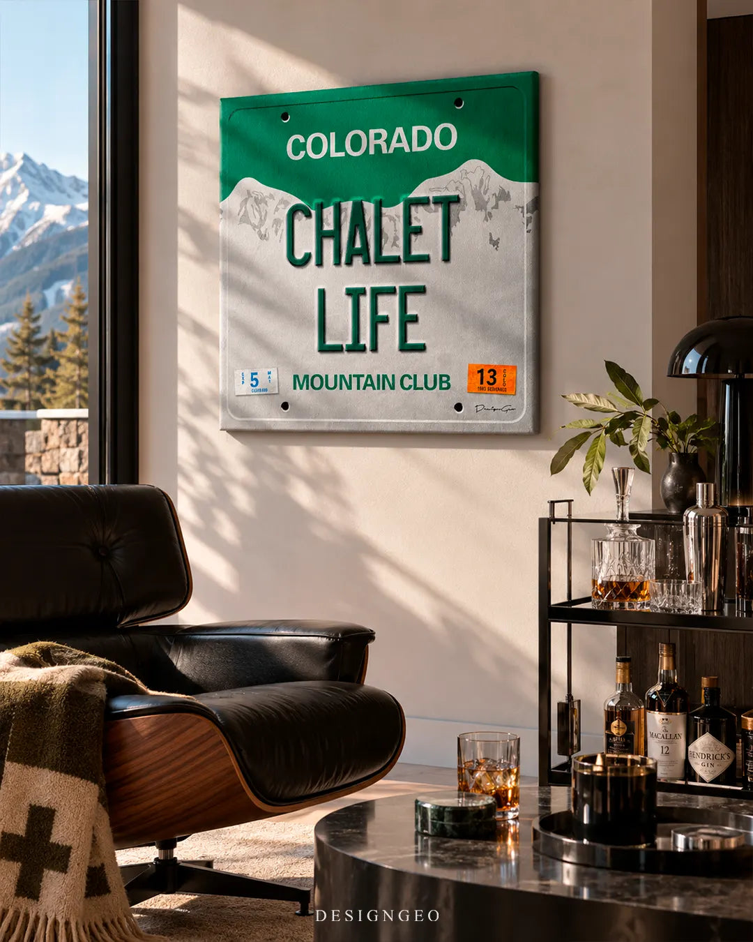 Luxury alpine lounge featuring a square Colorado Chalet Life license plate canvas wall art mounted on a neutral wall above a black Eames chair and a bar cart with whiskey decanters