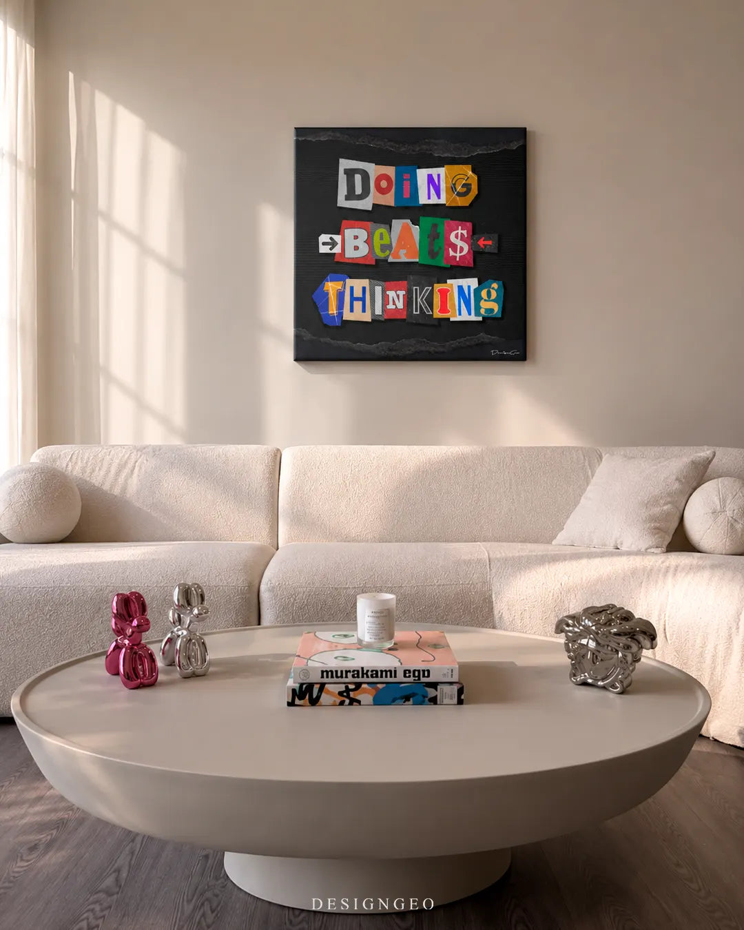 Square Doing Beats Thinking canvas print mounted on a beige wall above a off-white sofa and round coffee table with Murakami art books and balloon dog sculptures