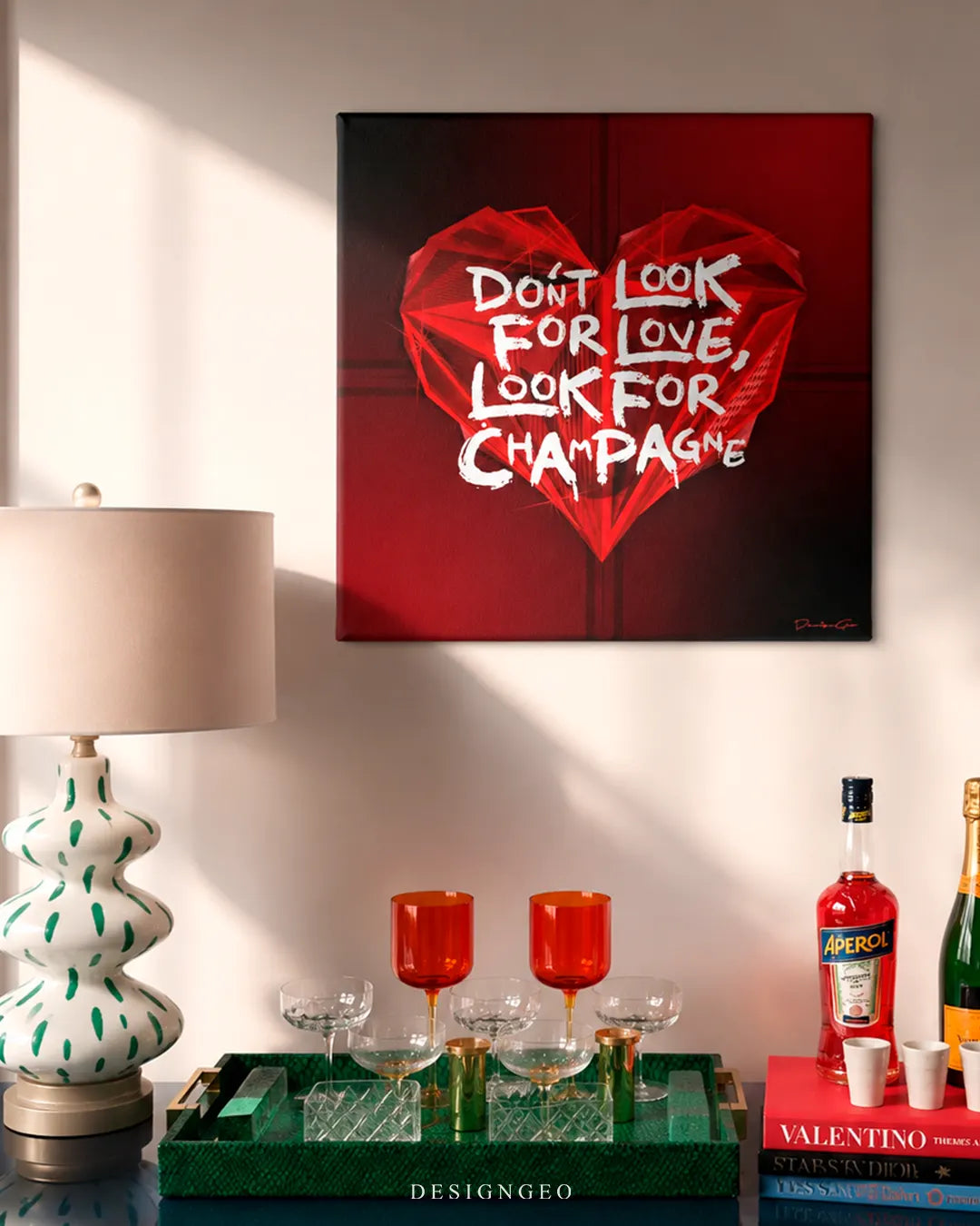 Square Don't Look For Love Look For Champagne canvas print mounted on a beige wall above a bar cart with crystal coupes, orange wine glasses, Aperol, and champagne bottles