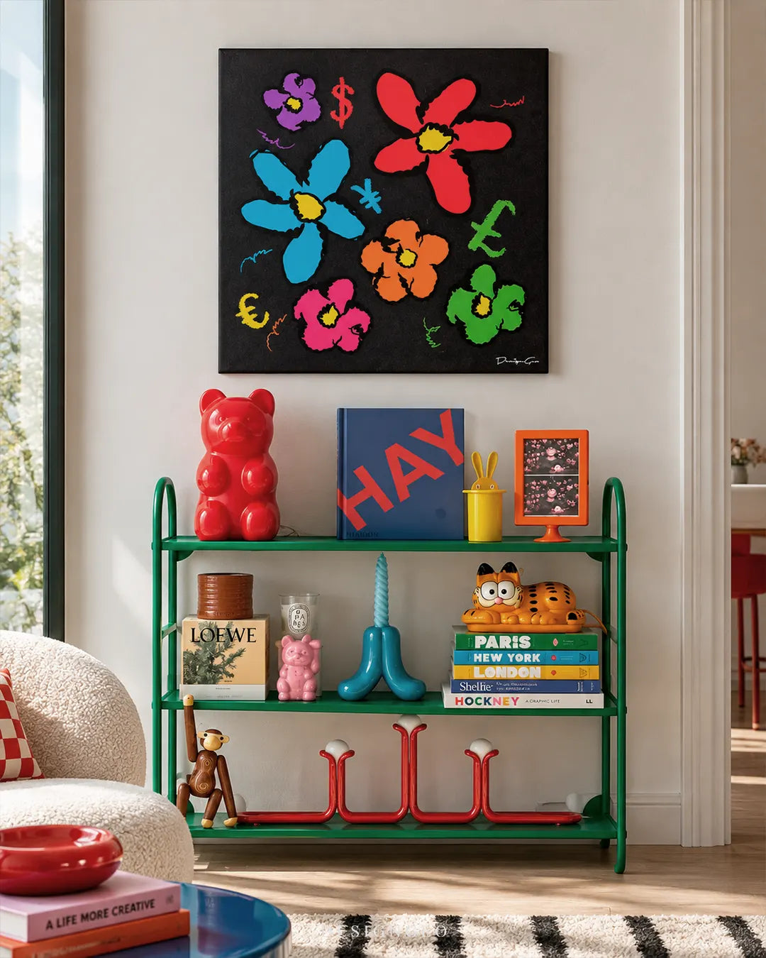 Square black canvas print featuring colorful pop art flowers and currency symbols mounted on a white wall above a green metal shelf styled with pop culture collectibles