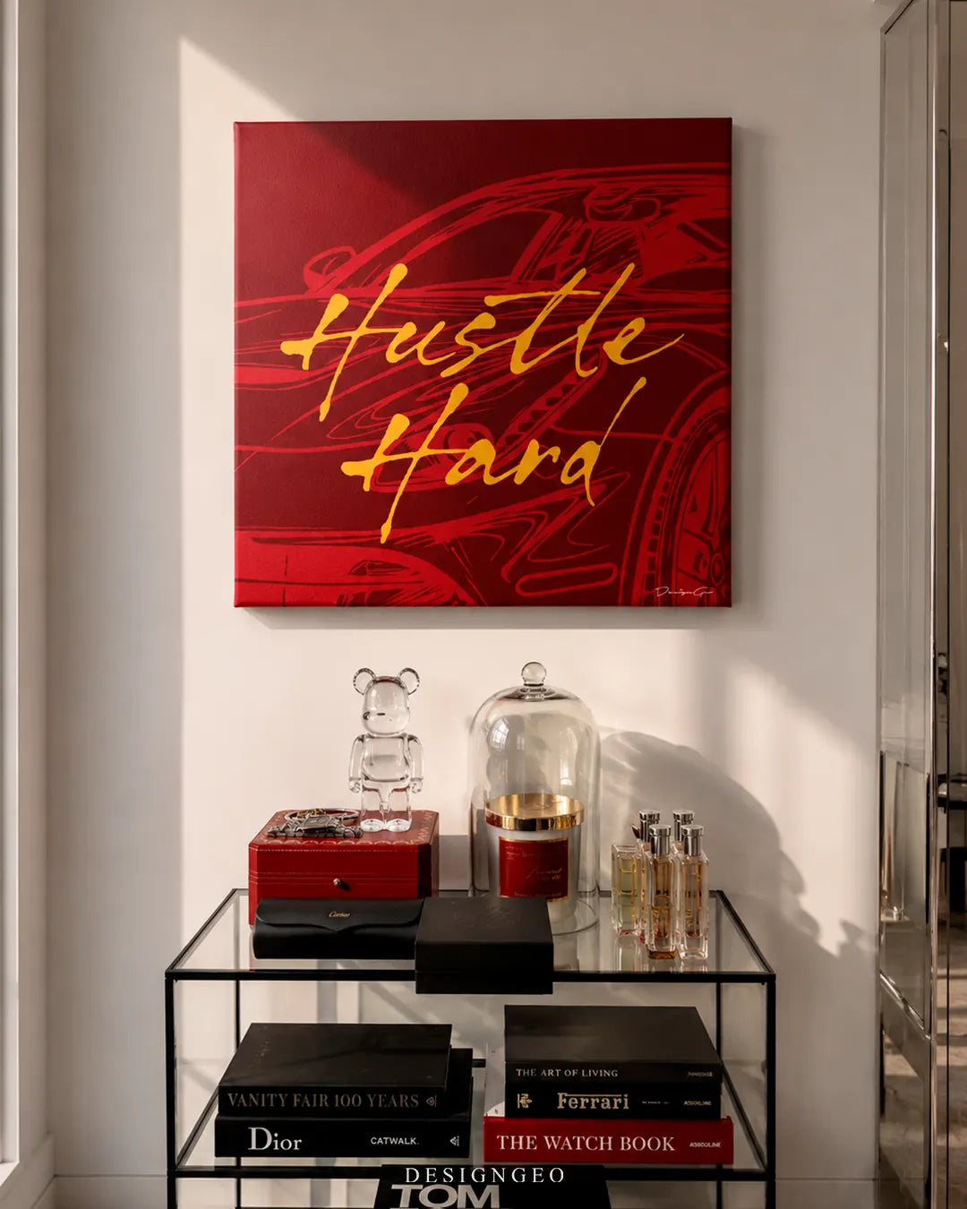 Square red and gold Hustle Hard motivational canvas print mounted on a white wall above a black glass display table with luxury perfumes, a clear Bearbrick figure, and luxury fashion books
