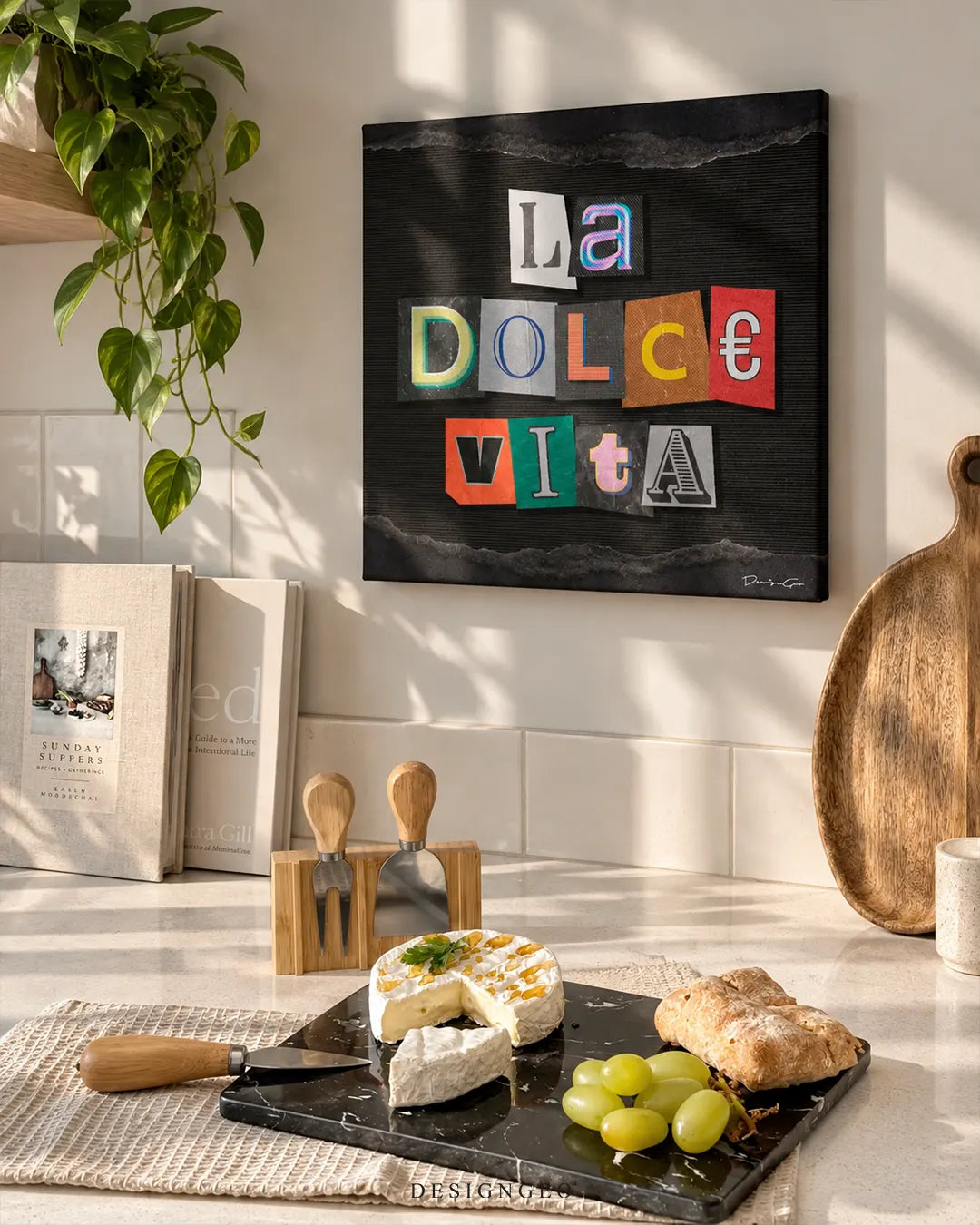 Square black canvas print featuring colorful cutout magazine letters spelling LA DOLCE VITA mounted on a beige tiled kitchen wall above a marble cheese board