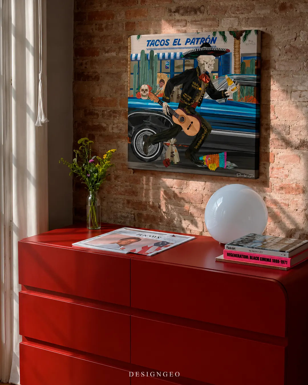 Square mariachi skeleton canvas print mounted on an exposed red brick wall above a sleek red credenza with a spherical white lamp, fresh flowers in a glass vase, and art magazines