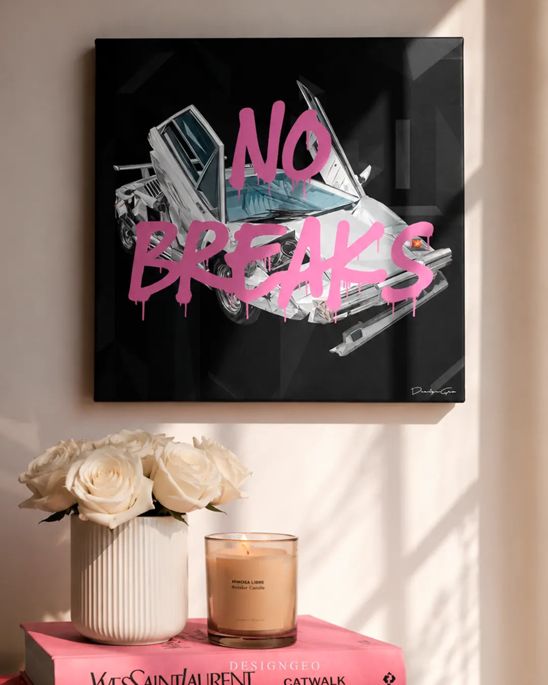 Square black No Breaks canvas print mounted on a beige wall above white roses, a lit candle, and a pink Yves Saint Laurent book