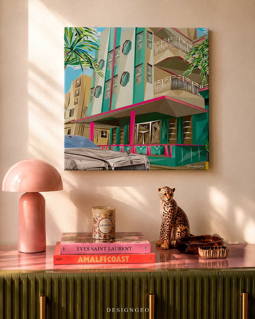 Square Ocean Drive Miami canvas print mounted on a beige wall above a fluted green console table with a pink dome lamp, leopard figurine, and stack of fashion books