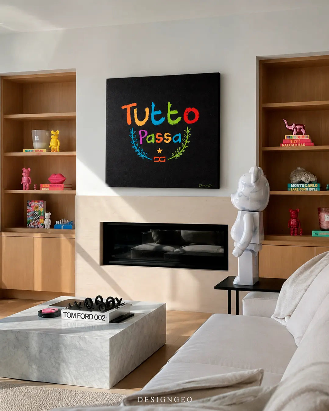 Square black Tutto Passa canvas print mounted above a modern fireplace in a luxury living room with built-in wooden shelves, a white Bearbrick statue, and a marble coffee table