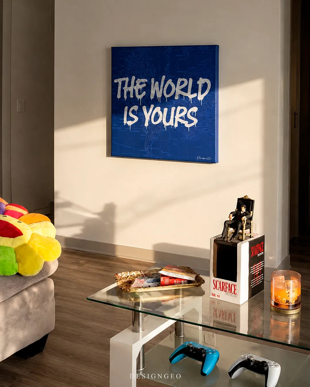 Square blue The World Is Yours canvas print mounted on a warm white wall above a glass coffee table with a mafia figure, gold tray, and gaming controllers
